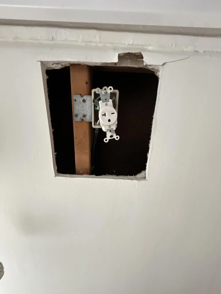 Square hole cut in white drywall revealing a 20-amp electrical outlet mounted to a metal box on a wood stud inside the wall cavity, indicating water damage investigation or remediation work