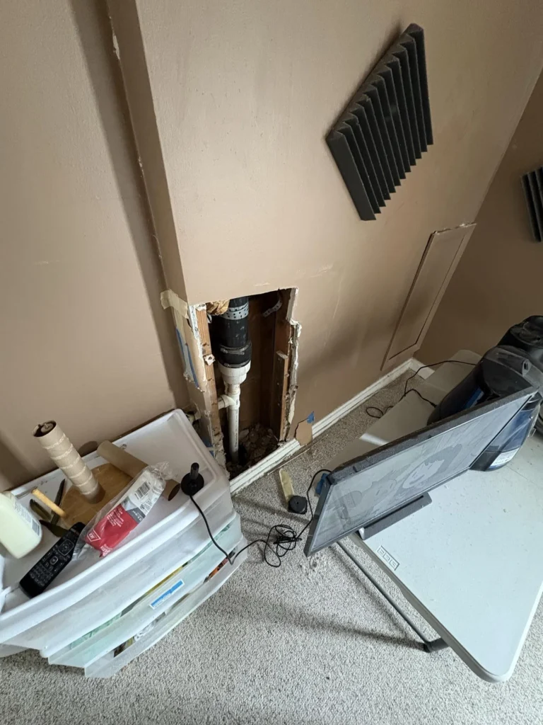 Interior wall with a vertical rectangular drywall cutout exposing a black ABS drain pipe and white PVC pipe inside the wall cavity, with an HVAC return vent nearby and items displaced on the floor during water damage investigation