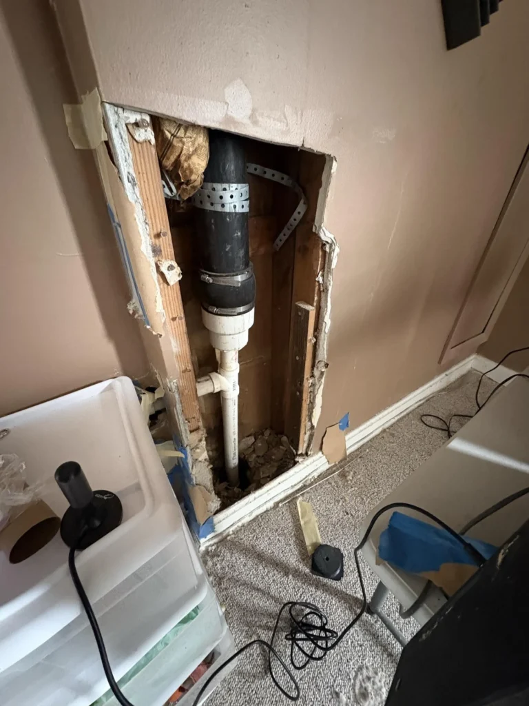 Close-up of a rectangular wall cutout showing a black ABS drain pipe connected to a white PVC pipe inside the wall cavity with metal hanger straps, surrounded by damaged drywall and wood framing during plumbing leak repair