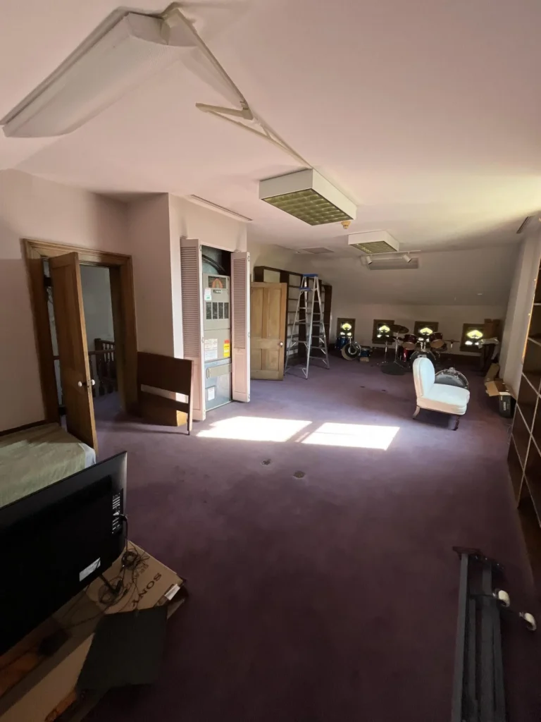 Large attic or ballroom space with purple carpet, collapsed and dangling ceiling tiles, an exposed air handler unit, a drum set, a white chaise lounge, wood bookshelves, and a ladder indicating water damage assessment