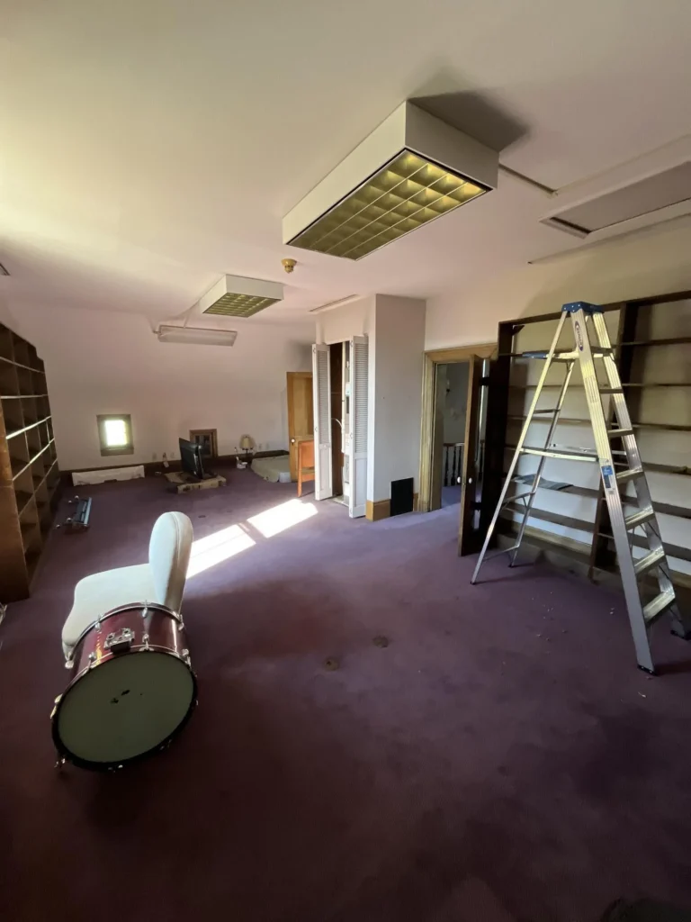 Large attic room with purple carpet, suspended ceiling tiles, an air handler unit in an open closet, a bass drum and chaise lounge, a stepladder, and empty bookshelves showing water damage impact