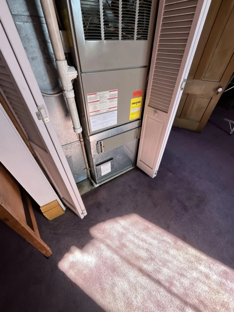 Close-up of a residential HVAC air handler unit inside an open louvered closet, showing the unit with EnergyGuide and warning labels, metal condensate drain piping, and an air filter housing
