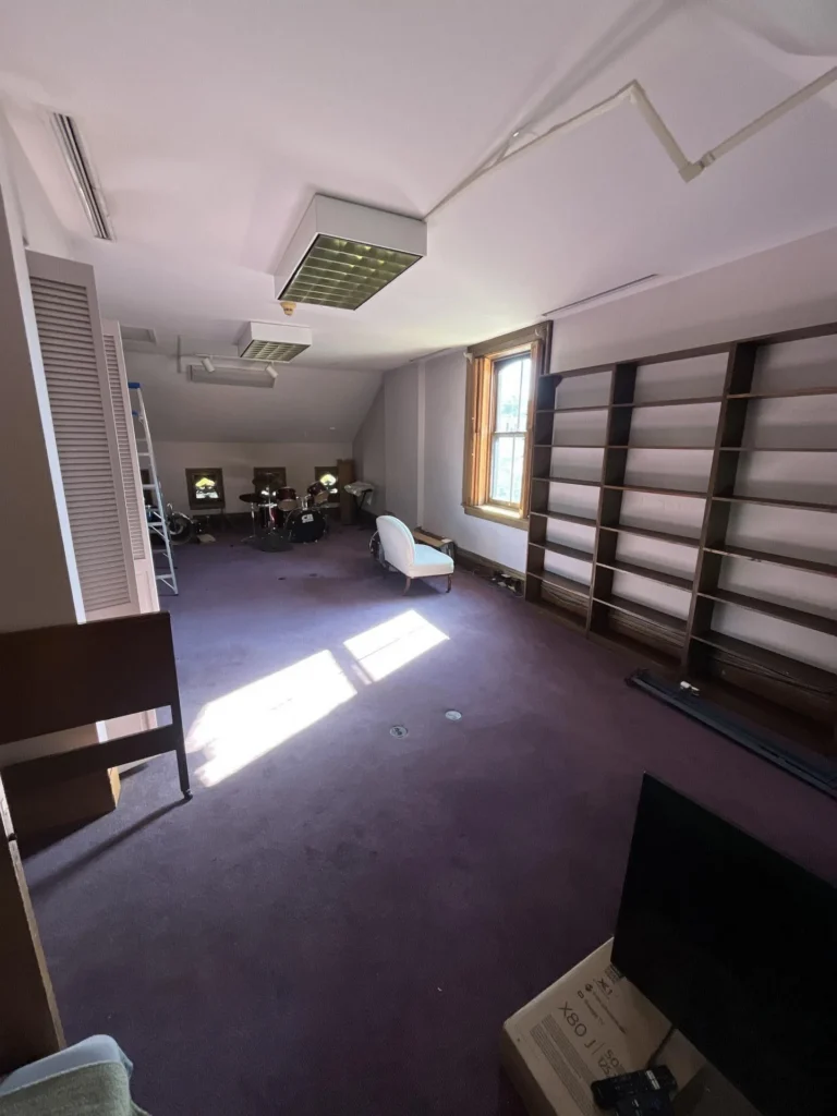 Attic room with purple carpet, large empty wood bookshelves on the right wall, a drum set and chaise lounge in the distance, a flat screen TV, and a ceiling with exposed ductwork from water damage