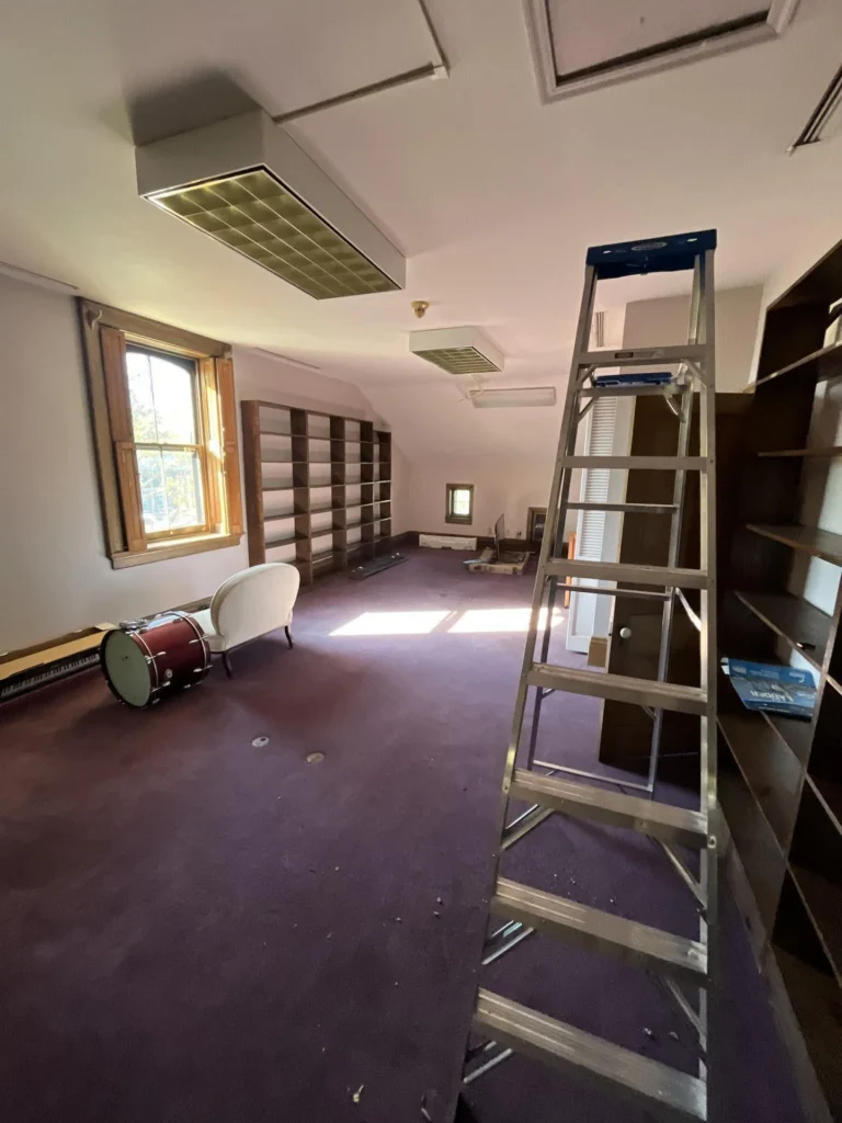 Spacious attic room with purple carpet, large empty wood bookshelves along the walls, a stepladder, a window with natural light, an air handler unit visible in a closet, and ceiling tile fixtures
