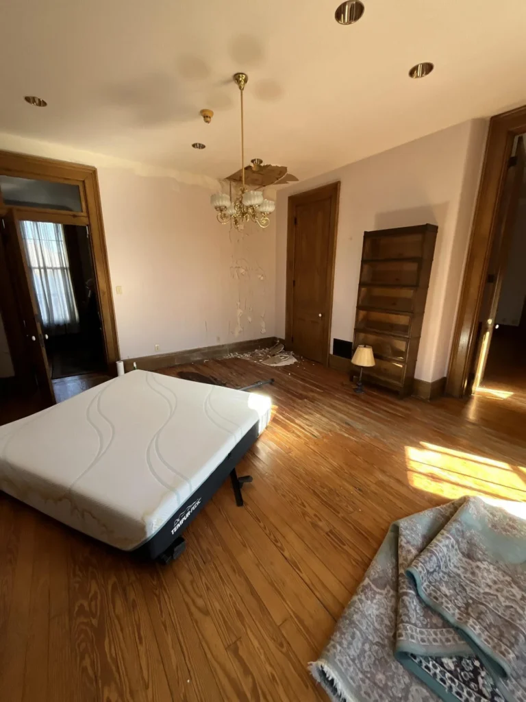 Water-damaged bedroom in a historic home with pink walls, a Tempur-Pedic mattress on the floor, a brass chandelier pulled partially from the ceiling with plaster debris fallen below, a bookshelf, and hardwood floors