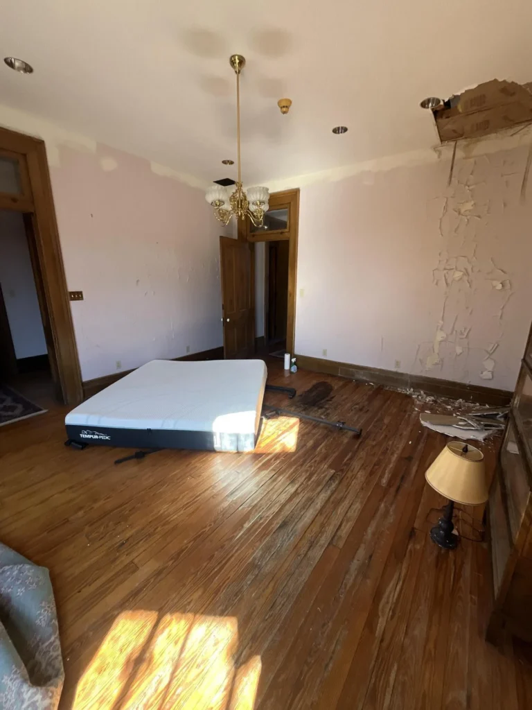 Historic home bedroom with water damage showing a Tempur-Pedic mattress on hardwood floor, a brass chandelier, a wall with severely peeling paint and a ceiling hole at upper right, a small lamp, and wood-trimmed doorways
