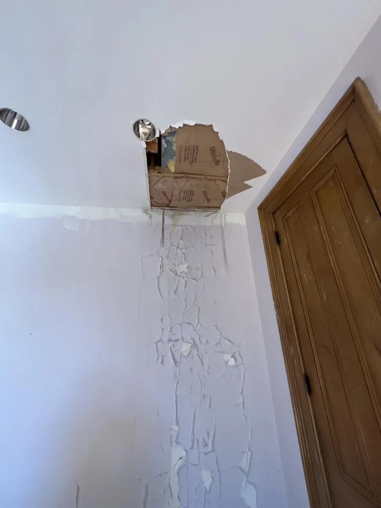 Bedroom wall and ceiling corner showing water damage with a large hole punched through the ceiling near a recessed light, exposing drywall backing, with extensive peeling paint running down the wall next to a wood-trimmed
