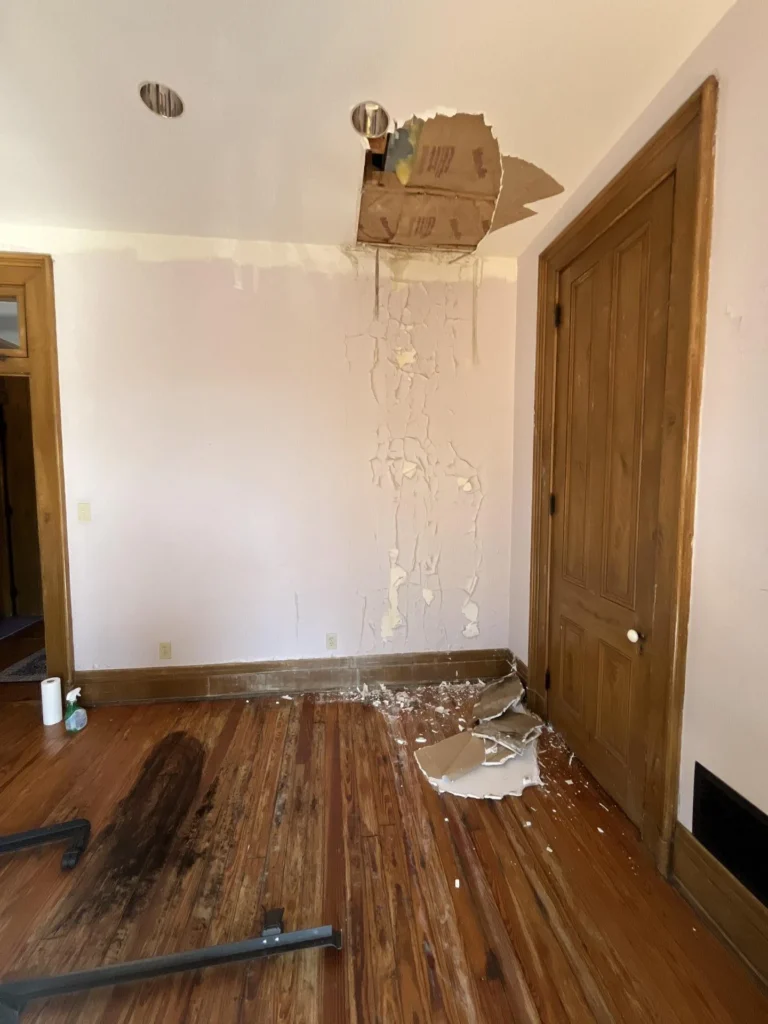 Bedroom with water damage showing a hole in the ceiling near a recessed light exposing drywall backing, extensive peeling paint streaking down the white wall, and large pieces of fallen drywall and plaster on the hardwood floor