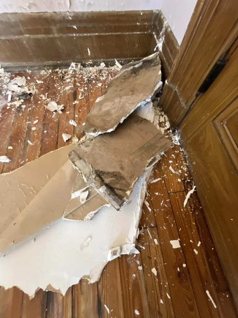 Water-damaged ceiling collapse with large chunks of drywall and plaster broken and fallen to the hardwood floor in the corner near a wood-trimmed door, with debris and dust scattered across the floor