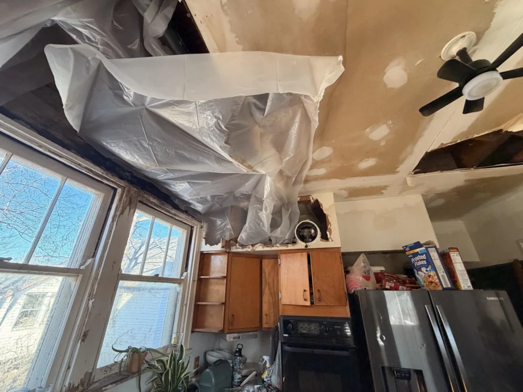Kitchen ceiling with extensive water damage showing exposed brown drywall backing, large sections of plaster fallen away, plastic sheeting hanging from upper cabinet area near a window, and a ceiling fan