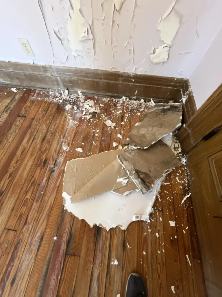 Bedroom corner with extensive water damage showing large sections of drywall fallen from the ceiling onto the hardwood floor, paint severely peeling from the wall below, and a wood baseboard visible